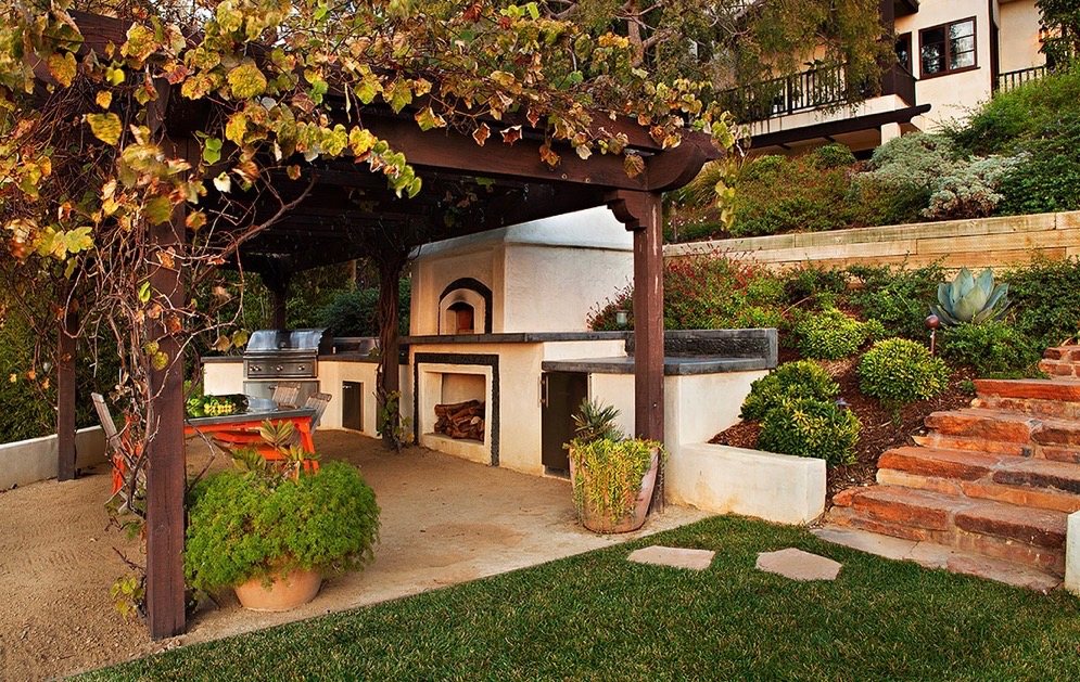Stucco outdoor kitchen with built-in grill and wood-fired pizza oven beneath a vine-covered timber pergola