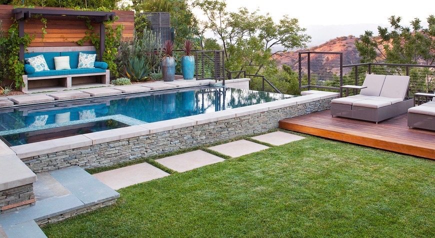 Hardwood lounge deck and stacked-stone wall beside a raised pool with built-in bench seating