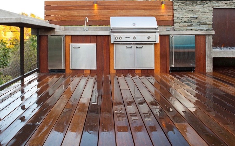 Deck-top outdoor kitchen with stainless grill, refrigerators, and concrete counters against a wood privacy wall