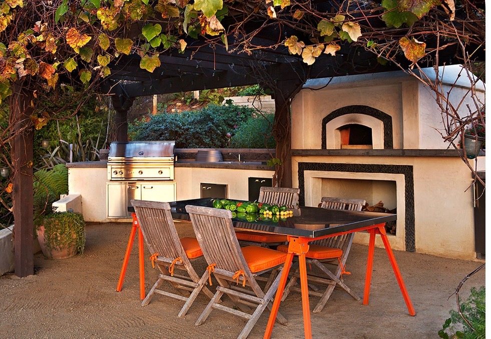 Outdoor dining table beneath a timber pergola in front of a stucco kitchen and pizza oven