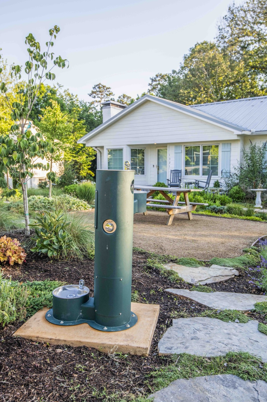 Green pedestal drinking fountain set in a planted garden with stone paving