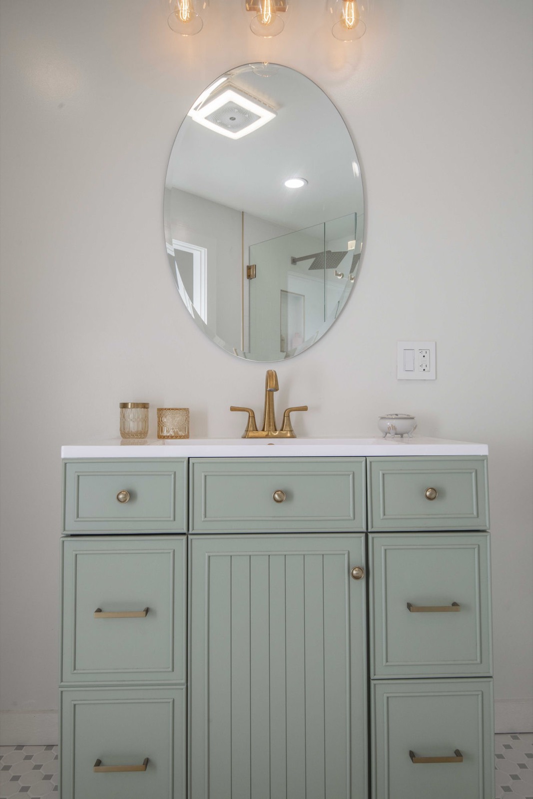 Sage-green vanity with brass fixtures, an oval mirror, and patterned floor tile