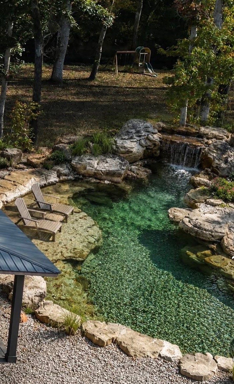 Boulder-edged natural pool with a small waterfall and lounge chairs on a rock ledge in a wooded yard