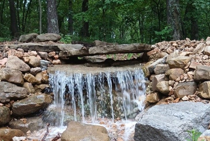 Water falling over a flat stone ledge between stacked boulders in a wooded setting